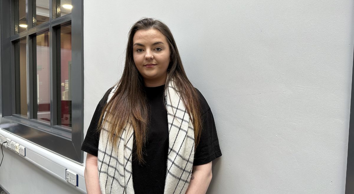 Woman with long brown hair wearing a short sleeved black top and a white check scarf standing against a white wall with window in background